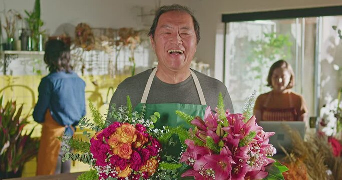 One happy mature Japanese small business owner of flower shop holding bouquets and smiling to camera standing inside retail store