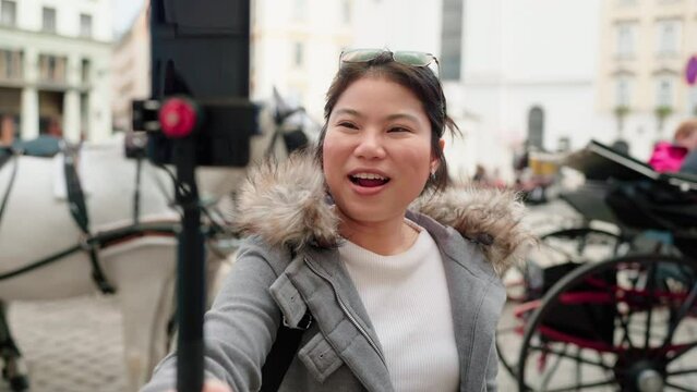 Young casual woman using smartphone to record journey with smile. Vienna Austria tour asian girl traveller wear stylish cloth with hand gesture waving to internet audience. Influencer vlogging concept