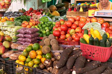 woman at market stall selling tomatoes, cassava, beans, corn on the cob, fruits and various sauces