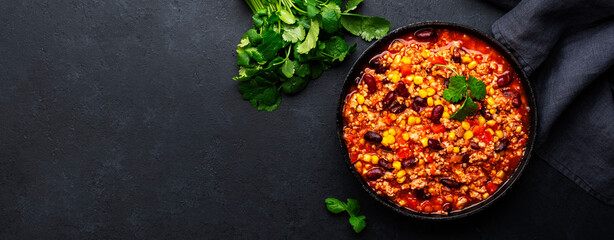 Chili con carne with beef, red beans, paprika, corn and hot peppers in tomato sauce, spicy tex-mex dish in a cooking pot, black table background, top view banner