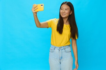 Asian woman holding her phone and looking at the screen taking pictures of herself talking on a video call with a yellow case on a blue background wearing a yellow T-shirt smiling with teeth 