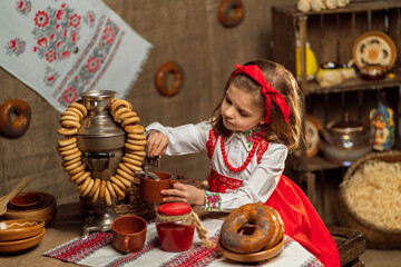 Little girl in folk dress pours tea from samovar while sitting in folk decor room. Shrovedite 