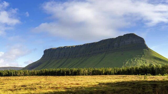 Time lapse of rural farming landscape with grass field and distant coniferous forest with moving clouds at Benbulben mountain on sunny day in county Sligo in Ireland.