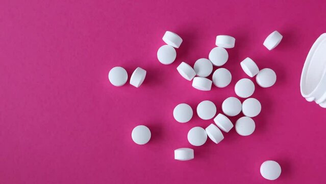 Round white pills spill out of the jar of pills on a pink background