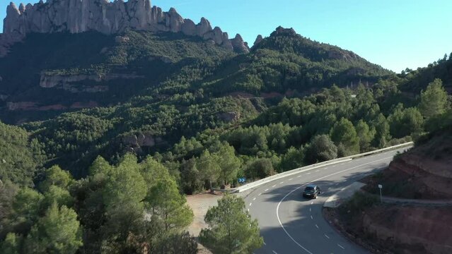 Cinematic aerial shot of old black car entering turn on mountain road with montserrat mountain in background