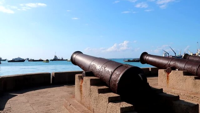 Panning shot of old rusted cannons with boats navigating in the port of Forodhani. Stone Town, Zanzibar