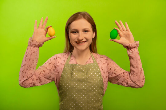 Happy smiling young woman housewife in green kitchen apron with polka dots and pink dress holding two multicolored painted dyed easter eggs isolated on green background.

Easter celebration concept.