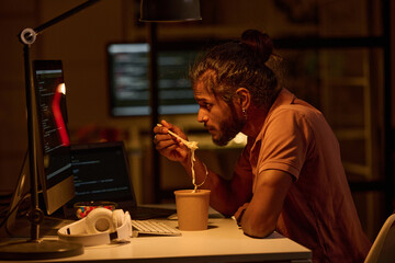 Young programmer examining codes on computer monitor and having lunch while working till late night in office