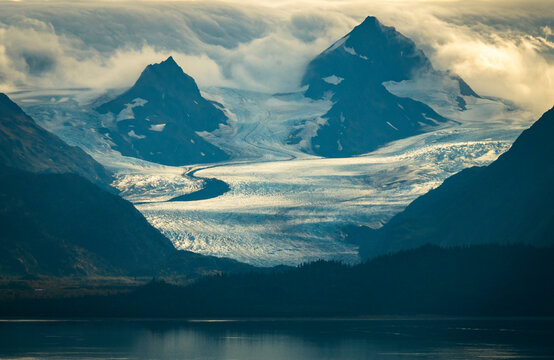 The morning sun rises over Kachemak Bay near Homer Alaska with clouds rolling over the mountain peaks and glaciers