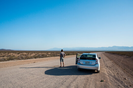 ARIZONA, USA. A man urinates on an empty dirt road leading into a vast desert. His hybrid car waits next to him.