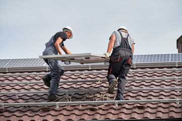 Men installers carrying photovoltaic solar modul on roof of house. Electricians in helmets...