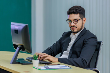 Millennial Asian Indian professional successful bearded male businessman in formal business suit and eyeglasses typing working with computer keyboard on work desk in multinational company office