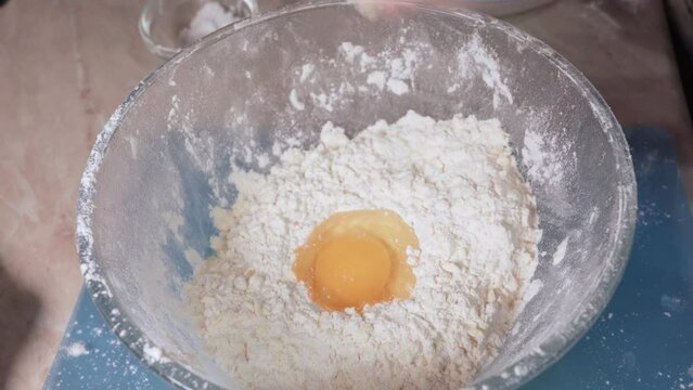 Women's hands add a chicken egg to the flour for making dough in a glass bowl.