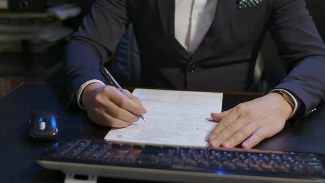 A man in a suit at a desk, near a computer, studying a form to compile (signing his resignation or filing his taxes). Panning rotating closeup shot.
