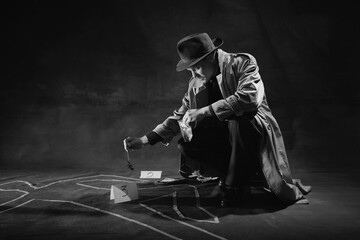 Black and white, noir photography. Man, detective in hat and trench coat near human drawn silhouette on floor, picking up clue. Concept of occupation, character, history. Retro style