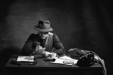 Black and white, noir photography. Serious man, professional detective in hat sitting at table and making crime notes. Investigation. Concept of occupation, character, history. Retro style