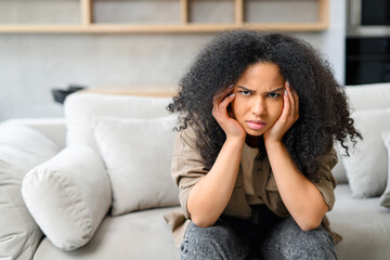 Fototapeta na wymiar Frustrated multiracial woman sitting on the sofa in grief, disappointed female feels despair, worrying, depression, loneliness, suffering from strong headache