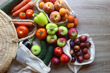 Straw bag and reusable fabric bags filled with various healthy fruit and vegetables. Wooden background, top view.