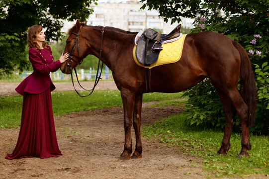 Pretty well dressed young woman stands next to cute brown horse and strokes it. Full horse in frame. Old fashioned picture of young lady in vintage burgundy suit and adult brown stallion on a walk