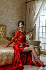 Pretty young woman with long legs in red dress sits on golden bed in luxury hotel room near window. Beautiful well dressed young lady is sitting in luxury palace. Luxury lifestyle, stylish jewelry