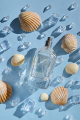 Glass bottle of perfume with seashells, ice cubes, water drops on the light blue background under sun rays. Smell of summer. Fresh  cooling perfumery. Ocean fresh smell. 