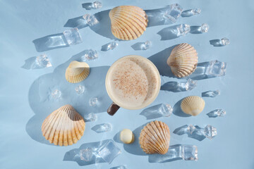 Cup of coffee with sea shells, ice, water drops on the blue background. Coffee latte with seashells in the middle of the table. Coffee break on  island. Happy day, morning. Enjoying vacation.