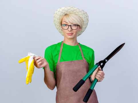 Young Gardener Woman With Short Hair In Apron And Hat Holding Hedge Clippers And Rubber Gloves Looking At Camera Smiling Confident Standing Over White Background