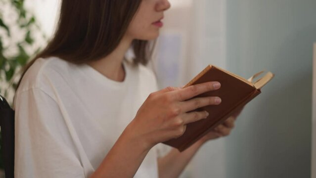 Focused woman with paralysis reads book with red cover flipping pages on blurred background. Person with disability sits in wheelchair enjoying hobby closeup