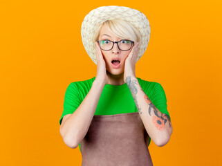 young gardener woman with short hair in apron and hat looking at camera being surprised and amazed standing over orange background