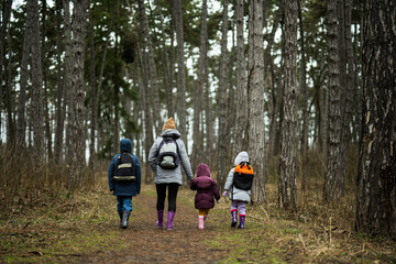 Back of mom and children with backpacks walking along the forest road after rain together.