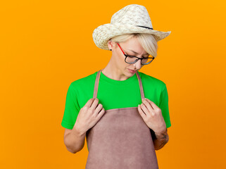 young gardener woman with short hair in apron and hat holding hands on chest looking down with sad expression standing over orange background