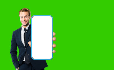 Excited smiling young business man in black suit, showing cell phone, mobile smartphone. Businessman hold cellphone with white screen, green chroma key background. App advertisement concept.