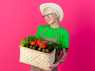 young gardener woman with short hair in apron and hat holding crate full of vegetables looking at camera with smile on face standing over pink background