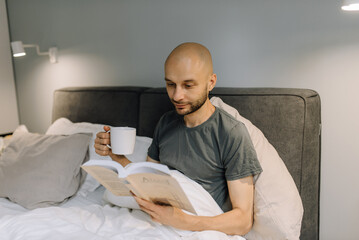 A man in bed reads a book and drinks hot tea or coffee