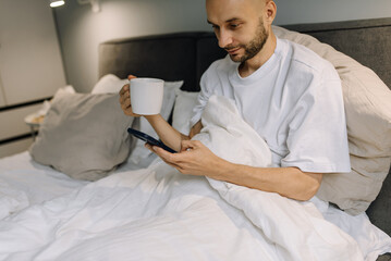 Smiling bearded man enjoying coffee in bed while holding phone. This is his morning ritual - starting the day with aromatic coffee and surfing the Internet