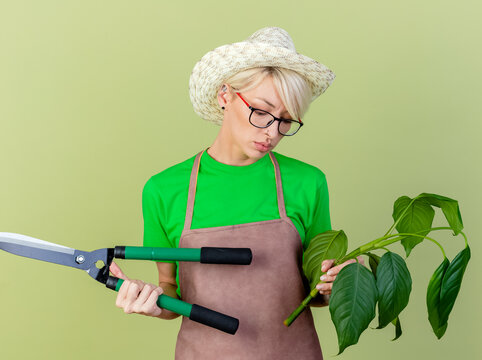 Young Gardener Woman With Short Hair In Apron And Hat Holding Plant And Hedge Clippers Looking Confused And Uncertain Standing Over Light Background