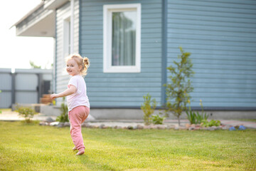 A small child runs on a green lawn. A beautiful and cheerful girl plays on the lawn near the house.