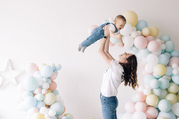 Mother plays with her son celebrating first birthday of baby boy among colorful balloons.