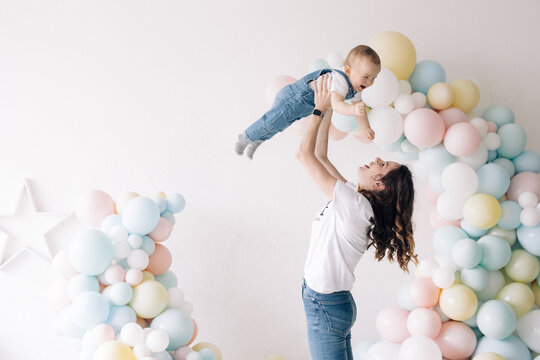 Mother plays with her son celebrating first birthday of baby boy among colorful balloons.