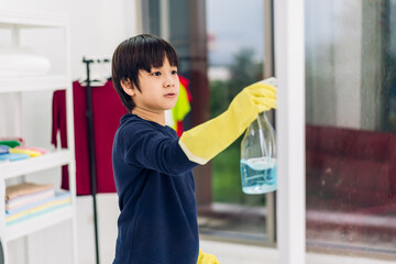 Kid little asian boy son having fun doing household cleaning and washing in protective gloves using a spray and rag while cleaning window in living room at home.housework and home cleaning concept