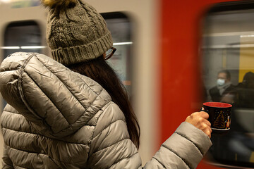 close de Mulher usando roupa de inverno, gorro e óculos segurando uma xicara com desenhos natalinos e na sua frente passa o trem do metro em velocidade.                       