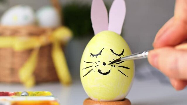Happy Easter. Womans hand draws a cute face with mustache and bunny ears on the eggshell of a painted egg with brush and paints. Decoration for the spring holiday. Preparation for the celebration.