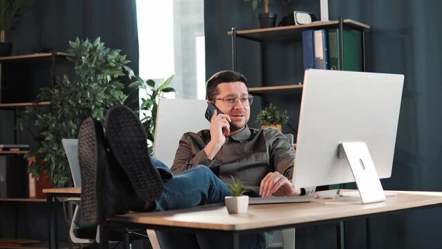 Calm relax free man put legs on table leaning on chair having mobile phone coversation calling enjoy day resting in modern office room looking at laptop screen, no stress, finish work, free time.