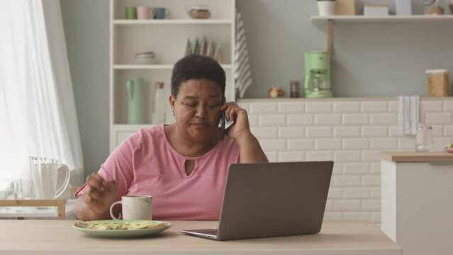 Waist up shot of mid aged African American woman talking on phone and using laptop while having breakfast in bright cozy kitchen