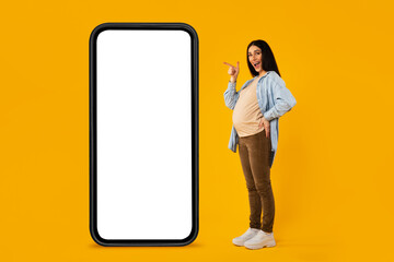 Happy pregnant woman pointing at big smartphone with white screen, standing on yellow studio background, mockup