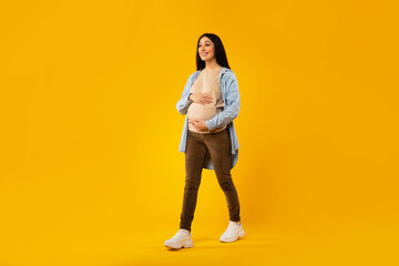 Fototapeta na wymiar Happy pregnancy concept. Pregnant woman walking and touching belly over yellow studio background, full length