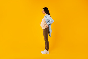 Fototapeta na wymiar Side view of pregnant lady standing isolated on yellow background and looking at free space, full length