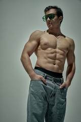 Handsome young man with strong muscular body shape posing in green sunglasses and jeans over grey studio background. Concept of man's beauty, sportive and healthy lifestyle, fashion