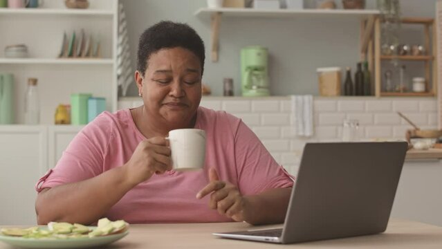 Waist up POV shot of happy mature African American woman speaking on camera while sitting in front of laptop at kitchen table