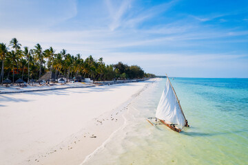 Relax on the white sand beach in Kiwengwa village on Zanzibar while admiring a Dhow catamaran sailboat gently gliding through the crystal-clear waters. - obrazy, fototapety, plakaty